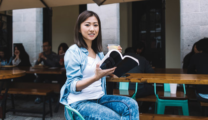 Asian smiling woman drinking refreshing beverage and reading book while looking at camera