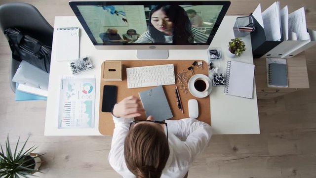 Flat lay of young beautiful female manager wearing white shirt and glasses sitting at desk, looking at computer screen and discussing different documents with Asian woman by video connection