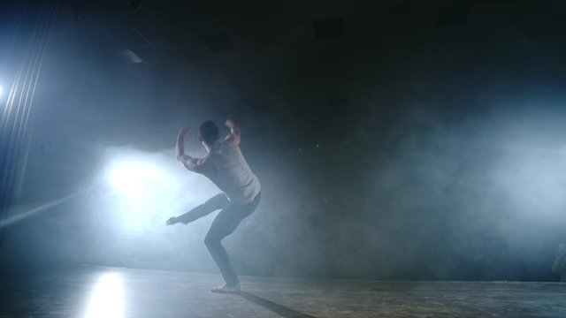 A modern ballet, a man performs jumps and spins in the light of spotlights and smoke on a dark background. Acrobatic choreography, rehearsal of the script of modern ballet