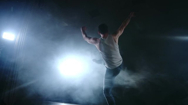 A modern ballet, a man performs jumps and spins in the light of spotlights and smoke on a dark background. Acrobatic choreography, rehearsal of the script of modern ballet