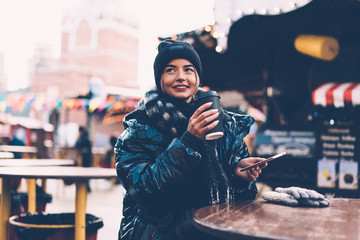 Modern happy woman with coffee cup using smartphone at Christmas market