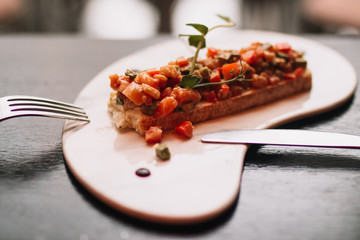 Bruschetta with salmon red fish, fresh vegetables and herbs on a white dish with a knife and fork on a dark background. Restaurant serving