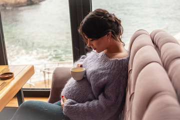 Young pregnant woman sit in cafe with beautiful view of window of the sea and mountains drink healthy tea . Female dressed warm woolen sweater and siting on the sofa.