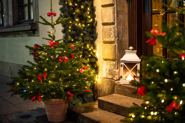 Christmas trees decorated with garland lightings and bowknots of red ribbon, vintage lantern outdoors on doorsteps in european old town house on christmas eve
