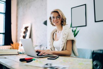 Portrait of cheerful hipster girl in classic optical glasses sitting at desktop in loft apartment...