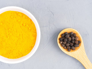 Turmeric in a white bowl and black pepper in wooden spoon on a gray concrete background. Healthy eating concept