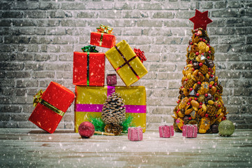 Mini Christmas tree or fir with gift boxes and a brick wall background with falling snowflakes. An image of a Christmas still life arranged on an old white oak table.