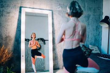 Healthy lifestyle exercise concept - Caucasian woman practices balance yoga asana Vrikshasana near lamp mirror, active girl in tracksuit doing tree pose for recreating soul and full minds indoors