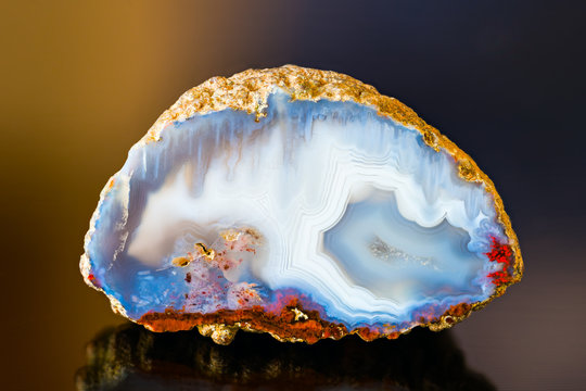 White blue agate gem cross section with reflection on dark colored background. One natural quartz chalcedony detail with crystals in a small geode. Red spots on smooth polished surface. Ice like look.
