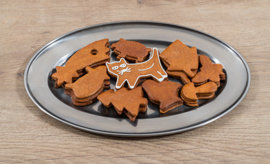 Cute white decorated gingerbread cat and mix of stacked cookies on silvery plate. Sweet holiday biscuits of varied shapes in shiny round platter on wooden background. Xmas, Children's or Cat Day.