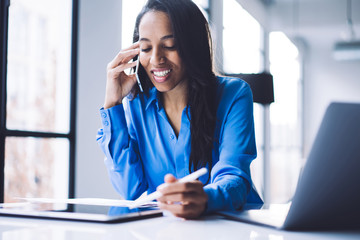 Positive black businesswoman talking on phone and using tablet at work