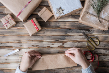 Woman packing handmade gifts made from kraft paper, christmas rope,  fir branches on the rustic wood planks background. DIY. Flat lay.