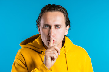 European handsome man holding finger on his lips over blue background. Gesture of shhh, secret, silence