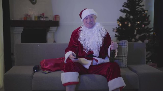 Portrait of positive Caucasian man in Santa Claus costume sitting on sofa with beer and watching TV at home. Senior man watching comedy show, resting at home after difficult working day.