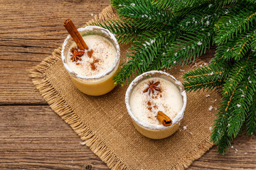 Eggnog cocktail in two glasses arranged with christmas decoration on old wooden table. Evergreen fer tree branch, artificial snow, sackcloth napkin