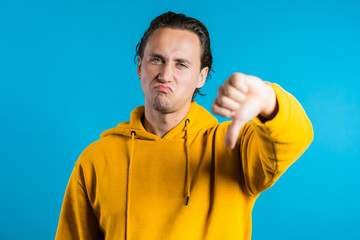 Young handsome man standing on blue studio background expressing discontent and showing thumb down...