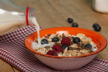 Pour the milk on the plate with oat flakes, dried fruits with blueberries and raspberries on wooden background. Healthy breakfast concept.