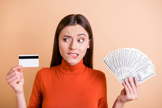 Photo Of Charming Beautiful Sad Upset Girlfriend Looking Suspiciously At Credit Card Without Understanding How It Works Holding Money Cash With Hands Inred Turtleneck Isolated Beige Color Background