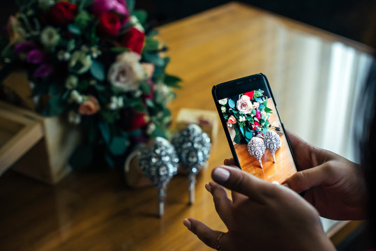 Woman hands taking photo of bright wedding flowers and luxury shoes with smartphone. Focus on bride's hand and display.