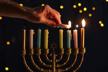 cropped view of man lighting up candles in menorah on black background with bokeh lights on Hanukkah