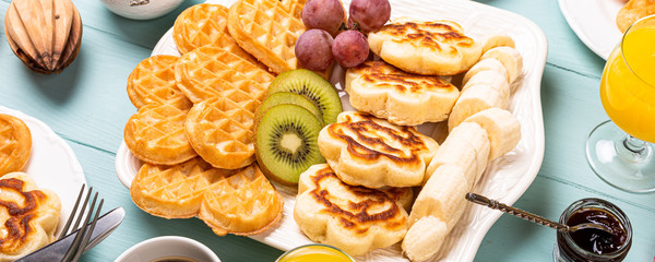 Healthy breakfast with fresh hot waffles hearts, pancakes flowers with berry jam and fruits on turquoise background, top view, flat lay. Food concept. Banner.