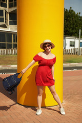 outdoor fashion portrait of a stylish young woman with a backpack in a short red dress, hat and sunglasses on colored background.
