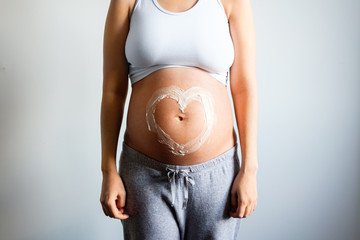 Future Mom posing with heart sign made from moisturising cream on naked belly , close up. Skin Care Moisturising and nutrition during pregnancy.