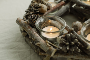 Close up view of decorative christmas wreath in scandinavian style made of cones and dried plants with light of candle on crumpled beige textile background.