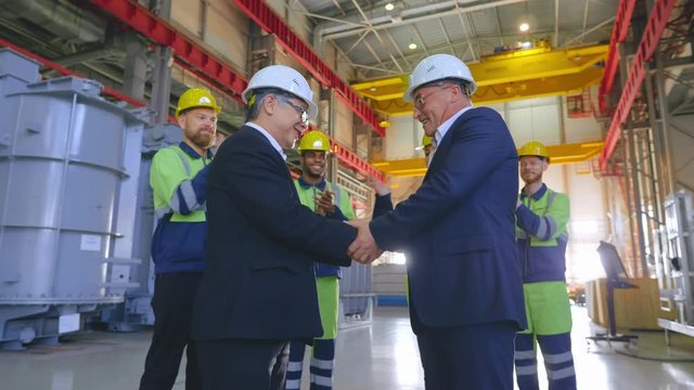 Engineers surrounded by workmen in hardhats greet each other with a handshake at a heavy industry factory. Successful deal of asian and caucasian contractors