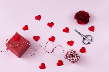 on a pink background is a red box with a gift, a ribbon for gift wrapping, a rosebud, scissors and red satin hearts