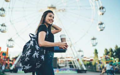 Smiling positive asian hipster girl in casual wear standing on wheel background with coffee to go, happy female student recreating in amusement park on weekends enjoying festivals and fan fairs