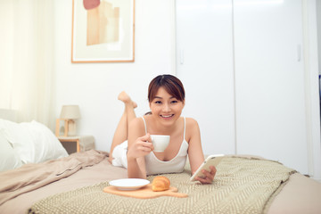 Obraz na płótnie Canvas Portrait of young Asian woman enjoying morning on bed while using mobile phone and drink tea/coffee