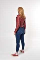 Fototapeta premium Full length portrait of a pretty blonde girl wearing red leather jacket denim jeans and sneakers. Standing pose, facing away from the camera, on a studio background.