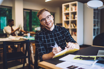 Portrait of happy male millennial smiling at camera during learning knowledge project in coworking space for students, smart casual hipster guy holding paper textbook for university education