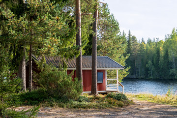 Fototapeta Red wooden finnish traditional cabins cottages in green pine forest near river. Rural architecture of northern Europe. Wooden houses in camping on sunny summer day obraz