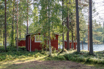 Red wooden finnish traditional cabins cottages in green pine forest near river. Rural architecture of northern Europe. Wooden houses in camping on sunny summer day