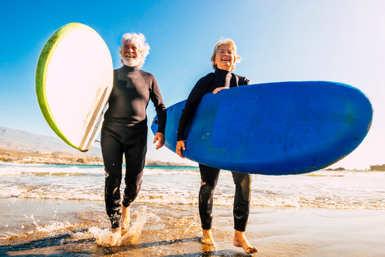 couple of seniors at the beach with black wetsuits holding a surftable ready to go surfing a the beach - active mature and retired people doing happy activity together in their vacations or freetime