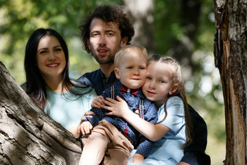 Family life. Portrait of parents and their children in park. Girl and boy sitting on a tree branch.