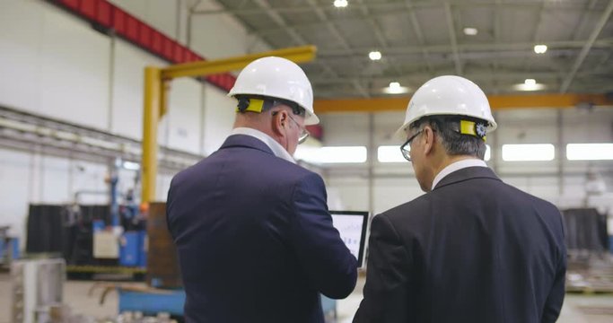 Businessmen engineers use tablet PC in industrial environment. People in formal suits walk with computer through workshop of modern factory
