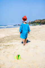 Little child in a Santa hat walks on a tropical beach.