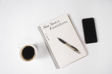 Phrase New Year's Resolutions in the notebook and pen. Cup of coffee and smart phone on table