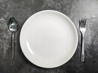 Empty White ceramic plate with fork and spoon on gray stone concrete table background. Copy space. Menu Recipe Concept