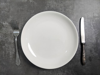 Empty White ceramic plate with fork and spoon on gray stone concrete table background. Copy space. Menu Recipe Concept