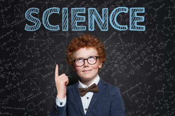 Smart confident kid boy with red hair pointing up at science inscription on chalkkboard