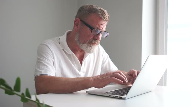 Tired elderly businessman works in the office with a laptop. The man takes off his glasses and wipes his eyes. Working weekdays, pensioner and computer