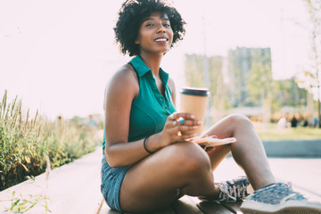 Portrait of charming african american woman enjoying free time outdoors on sunny summer day, attractive dark skinned hipster girl with curly chare resting with coffee to go on urban settings