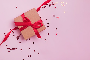 Gift box decorated with red bow on pink background with heart shape glitter confetti.