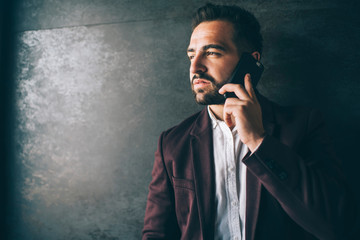 Handsome caucasian male entrepreneur in formal luxury jacket talking on mobile phone about business, confident man executive making smartphone call standing indoors on wall promotional background.