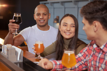 Handsome African man smiling to the camera, rising his beer glass enjoying drinking at the pub with his friends