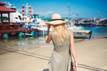 Fototapeta na wymiar Traveler blonde backpacker woman in straw hat back view walks along the coast at the pier fishing boats. Travel adventure in China.Tropical island Asia tourist. Summer holiday vacation journey concept